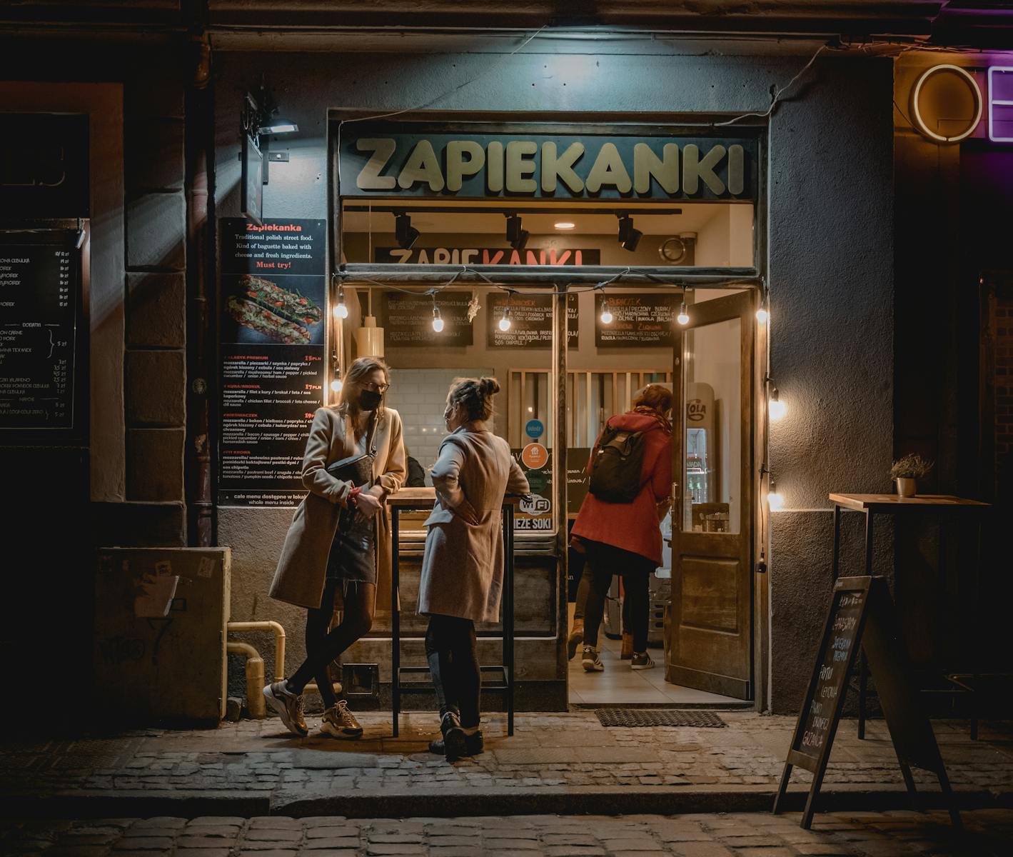 Two women chat outside Zapiekanki store at night in Poznań, capturing urban nightlife vibe.